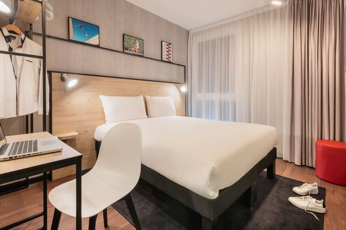 Et moderne hotelværelse med en seng, et skrivebord med en bærbar computer, en stol og en rød skammel.
