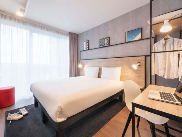 A modern hotel room with a large bed, a desk with a laptop, a red chair, and a wardrobe with clothes hanging. - Ibis Acheres St Germain En Laye (Acheres)
