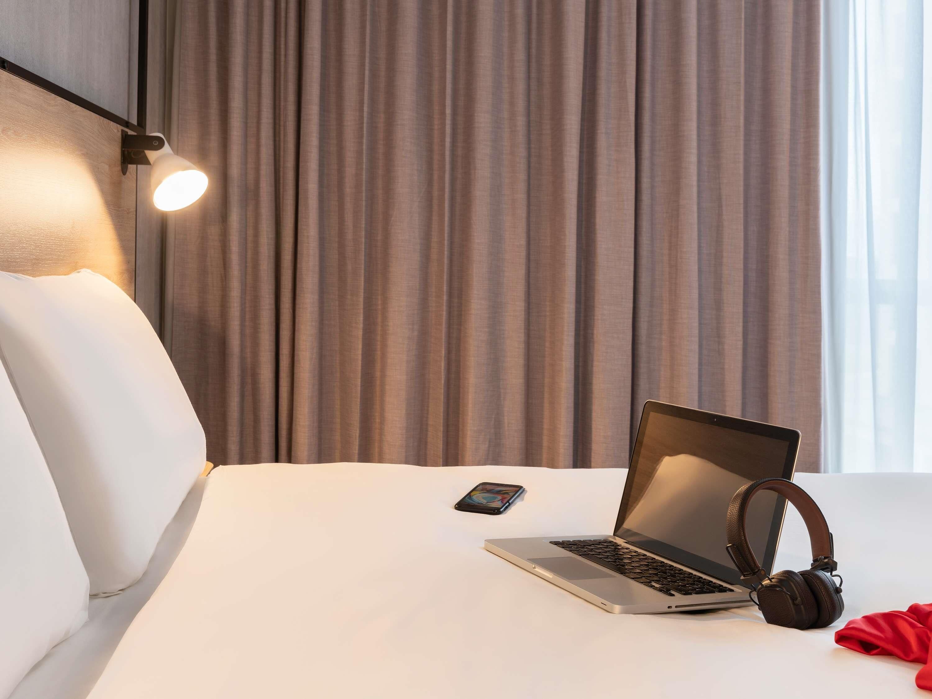 Une chambre d’hôtel avec un lit, un ordinateur portable, des écouteurs et un téléphone intelligent posés sur le lit.