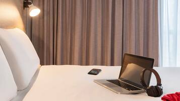 Une chambre d’hôtel avec un lit, un ordinateur portable, des écouteurs et un téléphone intelligent posés sur le lit.