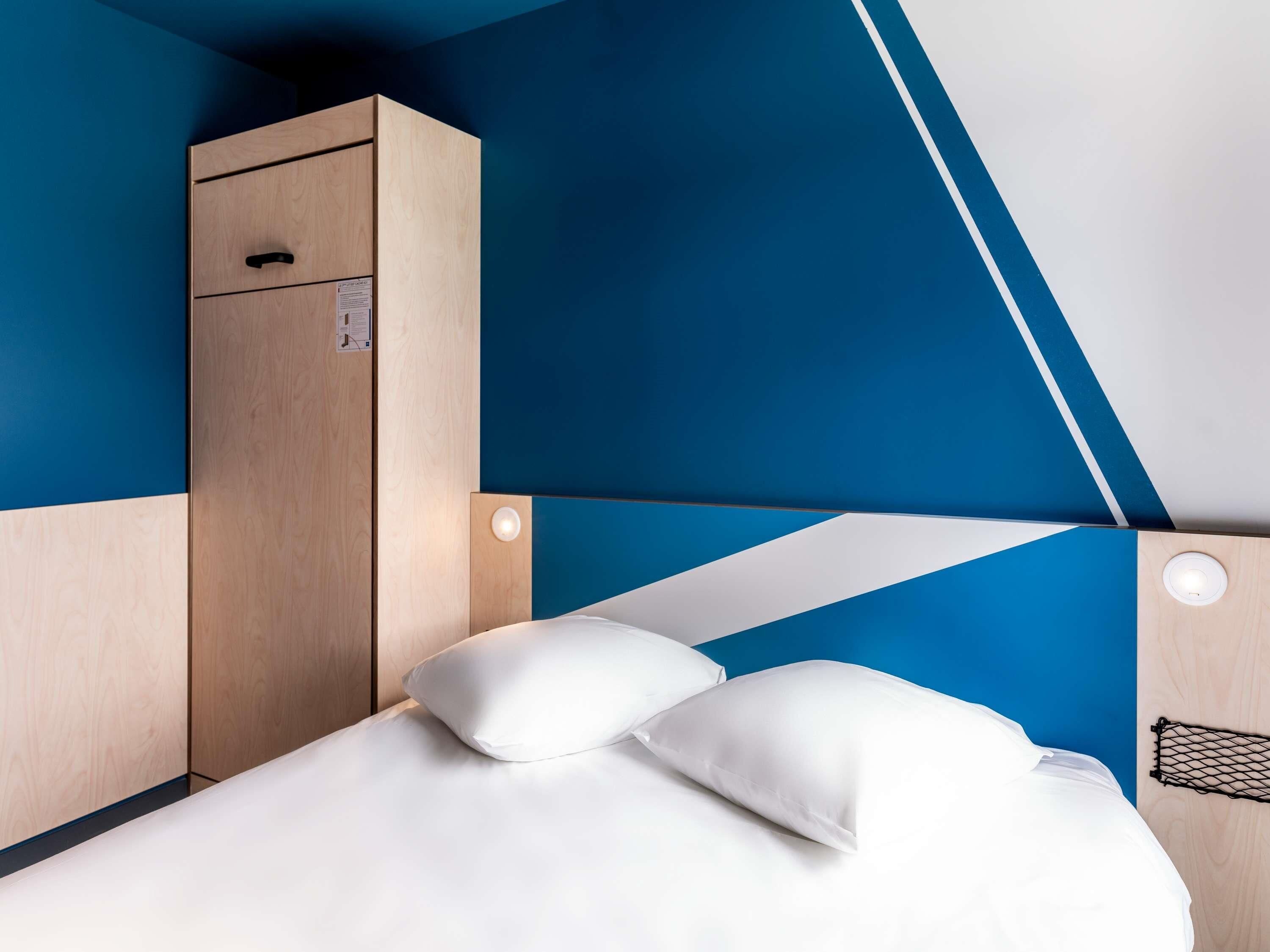 A modern hotel room with a bed, two pillows, a wooden wardrobe, and a blue accent wall.