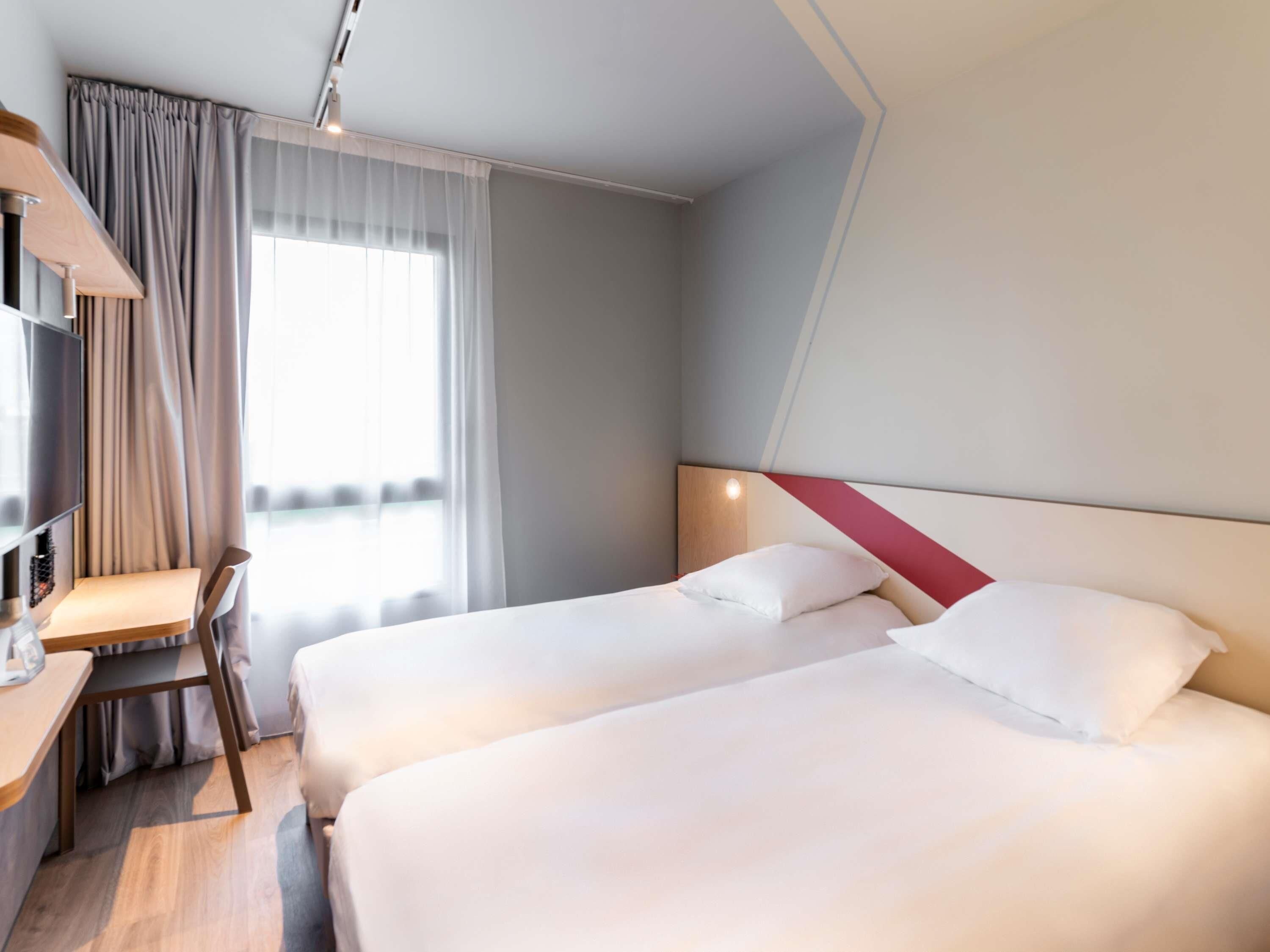 A hotel room with two beds, a desk, a TV, and a large window with curtains.