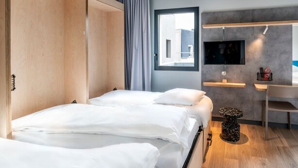 A modern hotel room with two beds, a desk, a TV, and a view of the city. - Ibis Budget Morlaix (Morlaix)