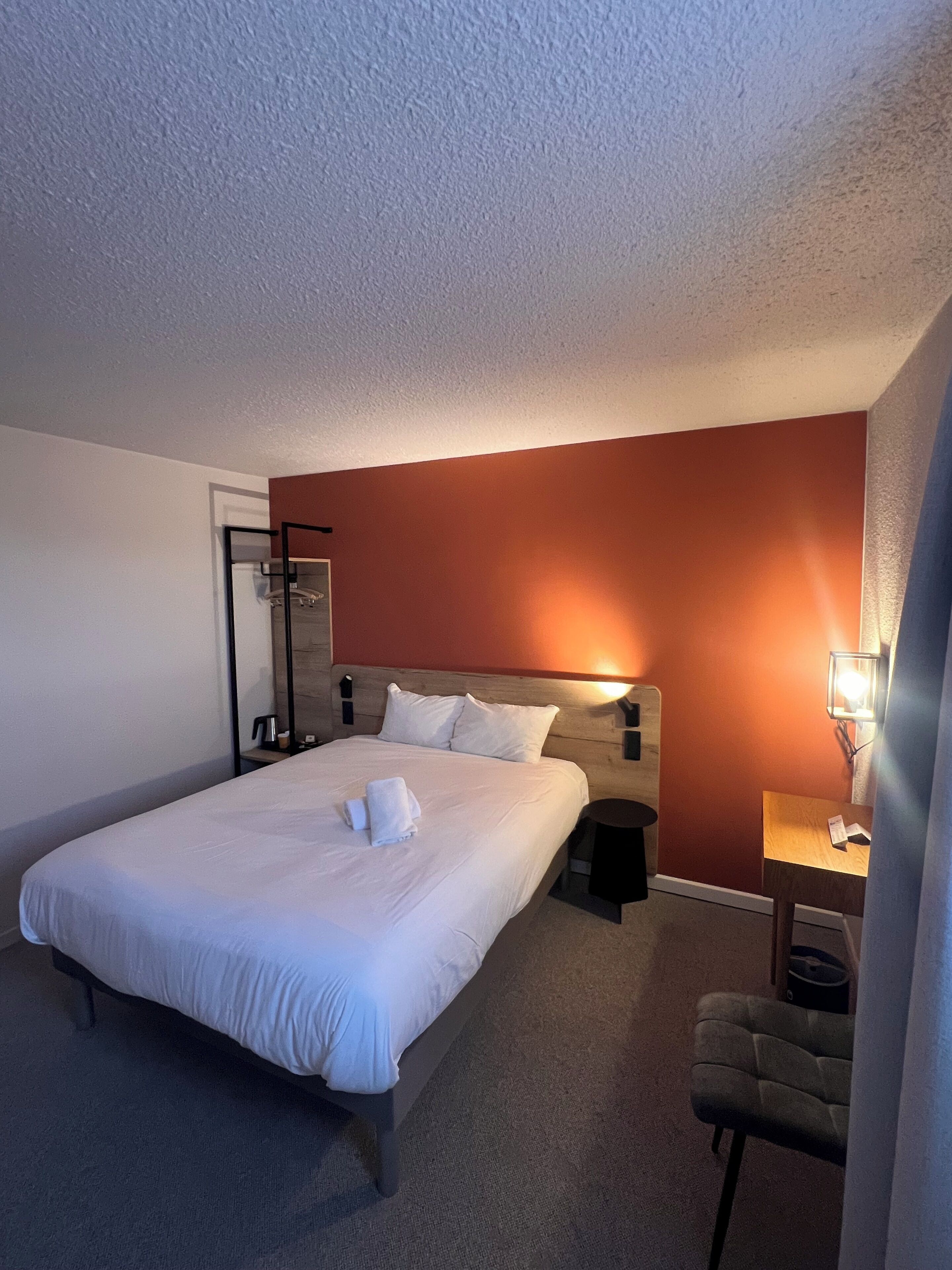 A hotel room with a large bed, a bedside table with a lamp, a small desk, and a chair.