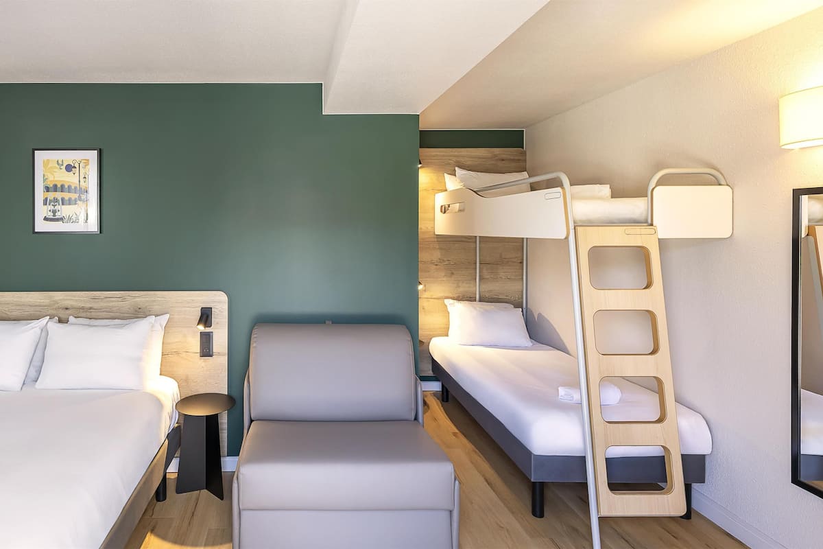 Une chambre d’hôtel avec un grand lit, un plus petit lit, une chaise, une échelle et une table de chevet avec une lampe.