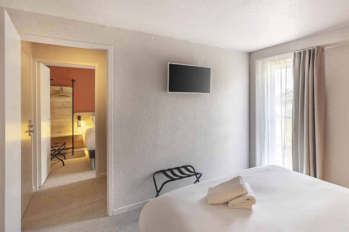 A hotel room with a bed, a TV mounted on the wall, a chair, and a window with curtains.