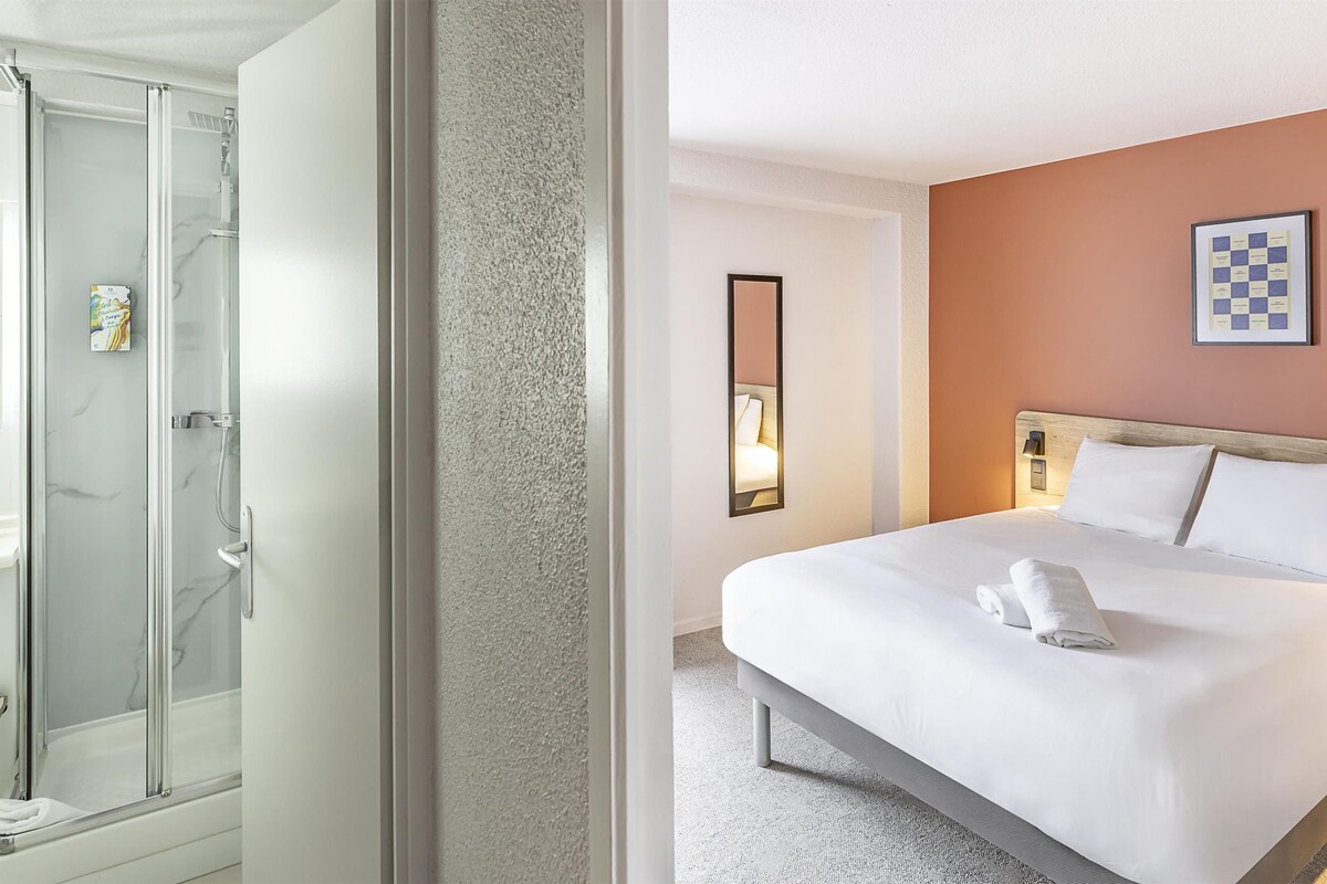 Habitación de hotel con ducha envuelta en vidrio, cama bien hecha con toalla enrollada y un tablero de ajedrez enmarcado en la pared.