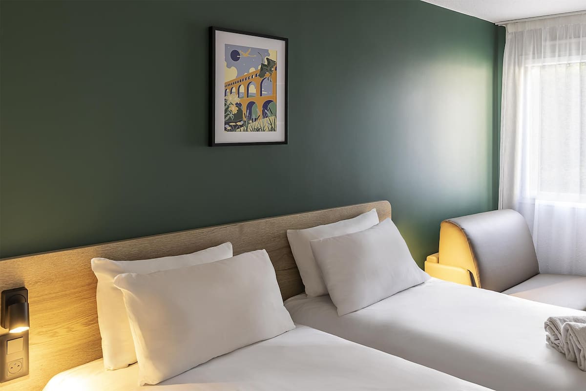 Une chambre d’hôtel avec un lit, une chaise, un tableau au mur et une fenêtre avec des rideaux.