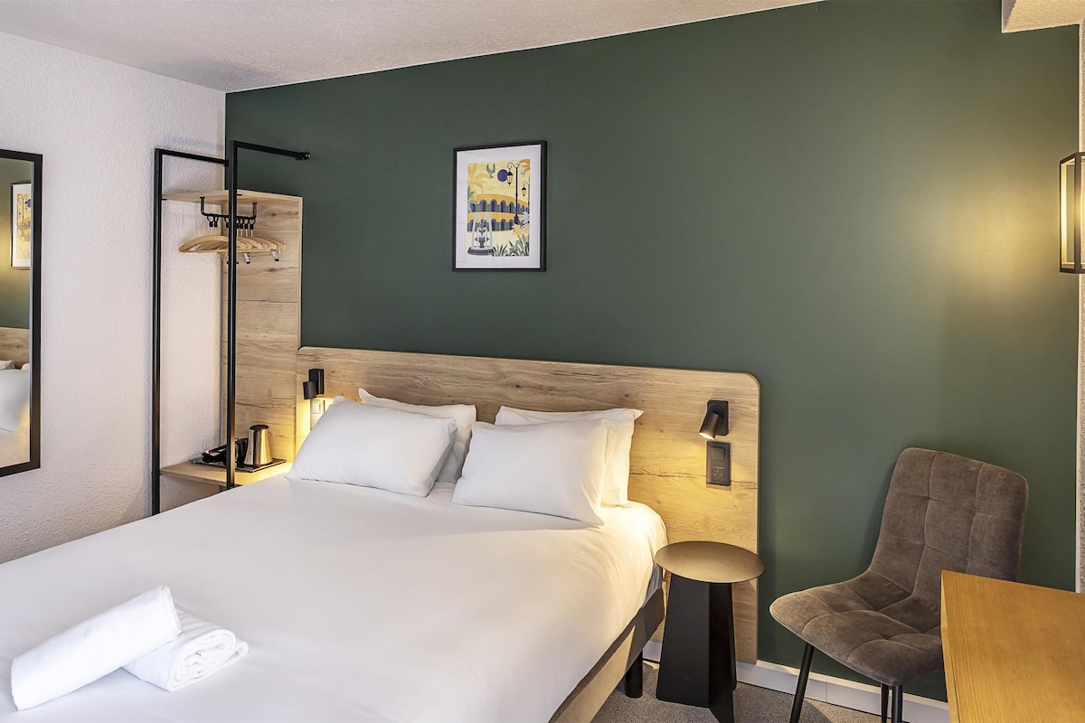 A hotel room with a large bed, a chair, a small table, a mirror, and a wall-mounted light fixture.