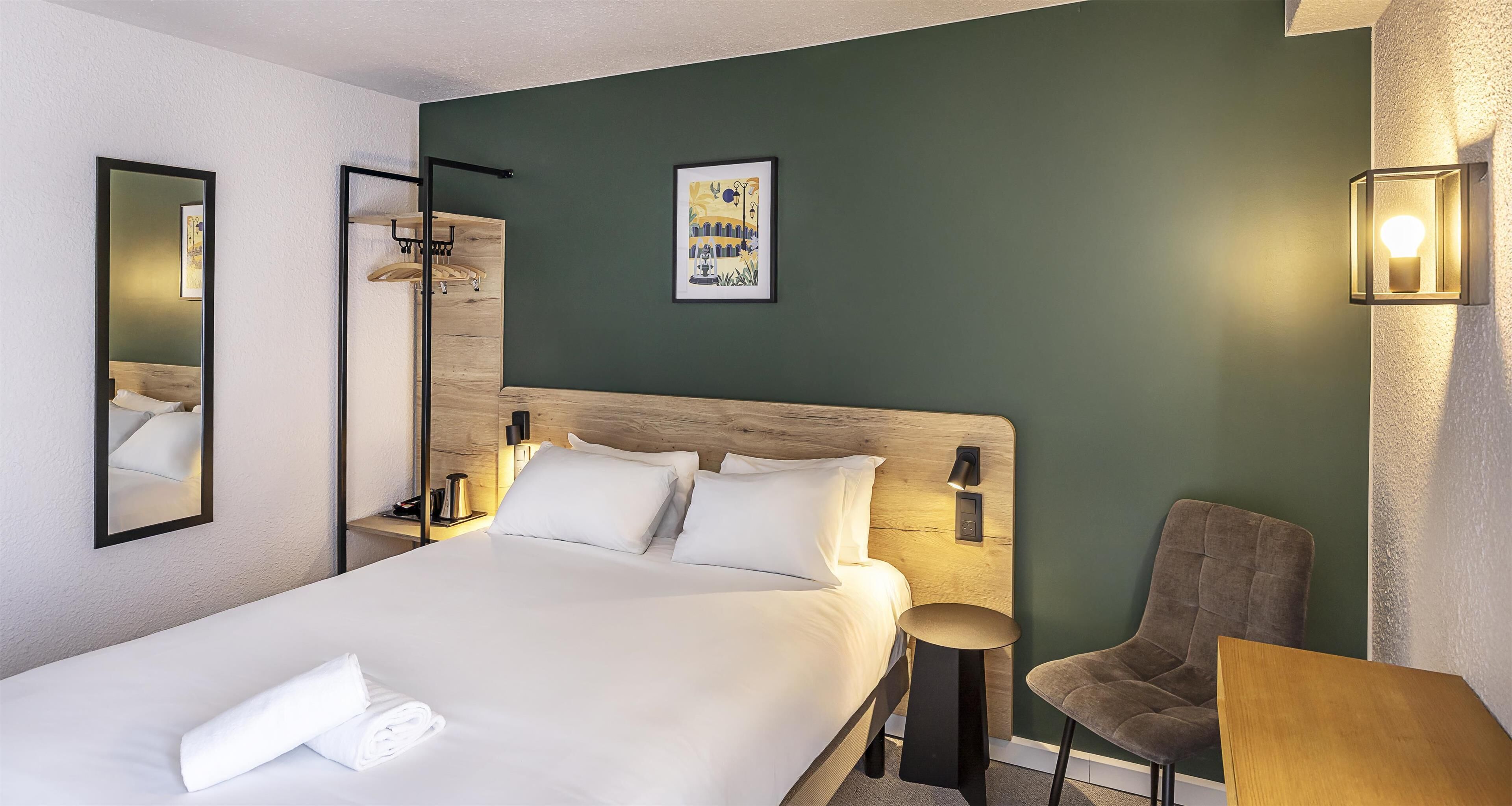 Une chambre d’hôtel avec un grand lit, une chaise, une petite table et un luminaire fixé au mur.