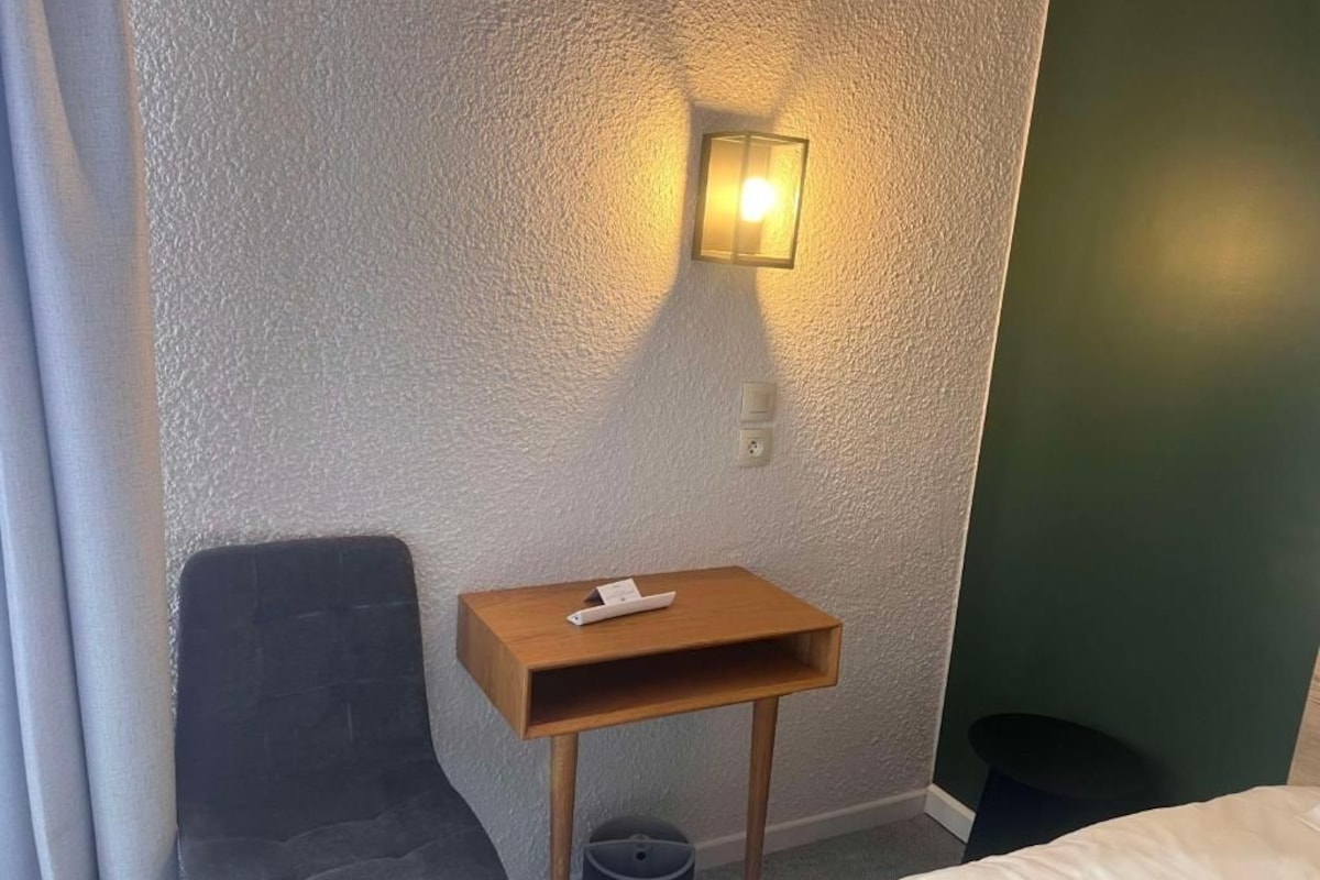 A hotel room with a single bed, a small wooden nightstand, a chair, a trash bin, and a wall-mounted light fixture.