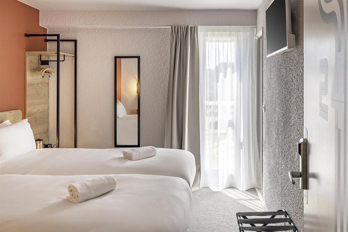 Une chambre d’hôtel avec deux lits, un grand miroir, une armoire et une vue sur la ville à travers des rideaux transparents.