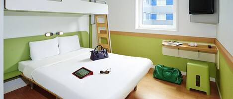 A hotel room with a bunk bed, a bedside table with a TV, a window with a view of buildings, and a green bag on the floor.
