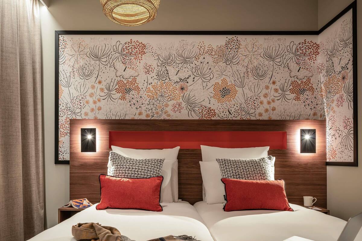 Une chambre d’hôtel avec un grand lit, deux oreillers, un ordinateur portable, une table de chevet et un mur recouvert d’un papier peint à motifs floraux.