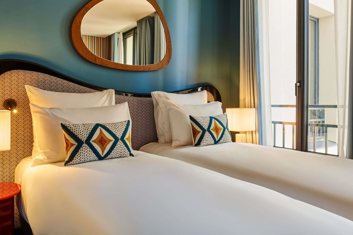 A hotel room with a large bed, two bedside tables, a round mirror, and a view of the outdoors.