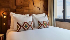 A hotel room with a wooden headboard, a large bed with white bedding, two patterned pillows, and a view of the city through a window. - Mercure Paris Butte Montmartre Basilique (Paris)