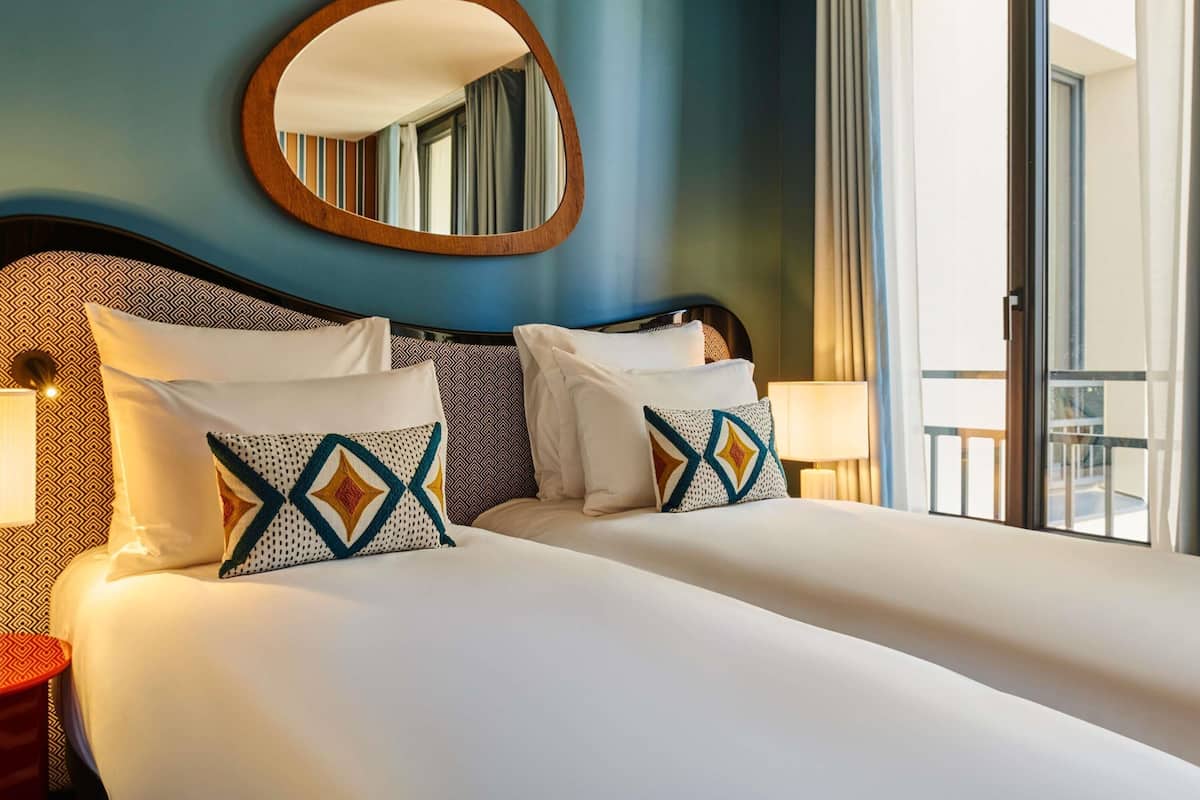 A hotel room with a large bed, two bedside tables, a round mirror, and a view of the outdoors.
