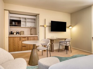 Habitación de hotel moderna con kitchenette, comedor con una mesa redonda y sillas, televisor de pantalla plana montado en la pared y cama con cubrecama estampado en verde y blanco.