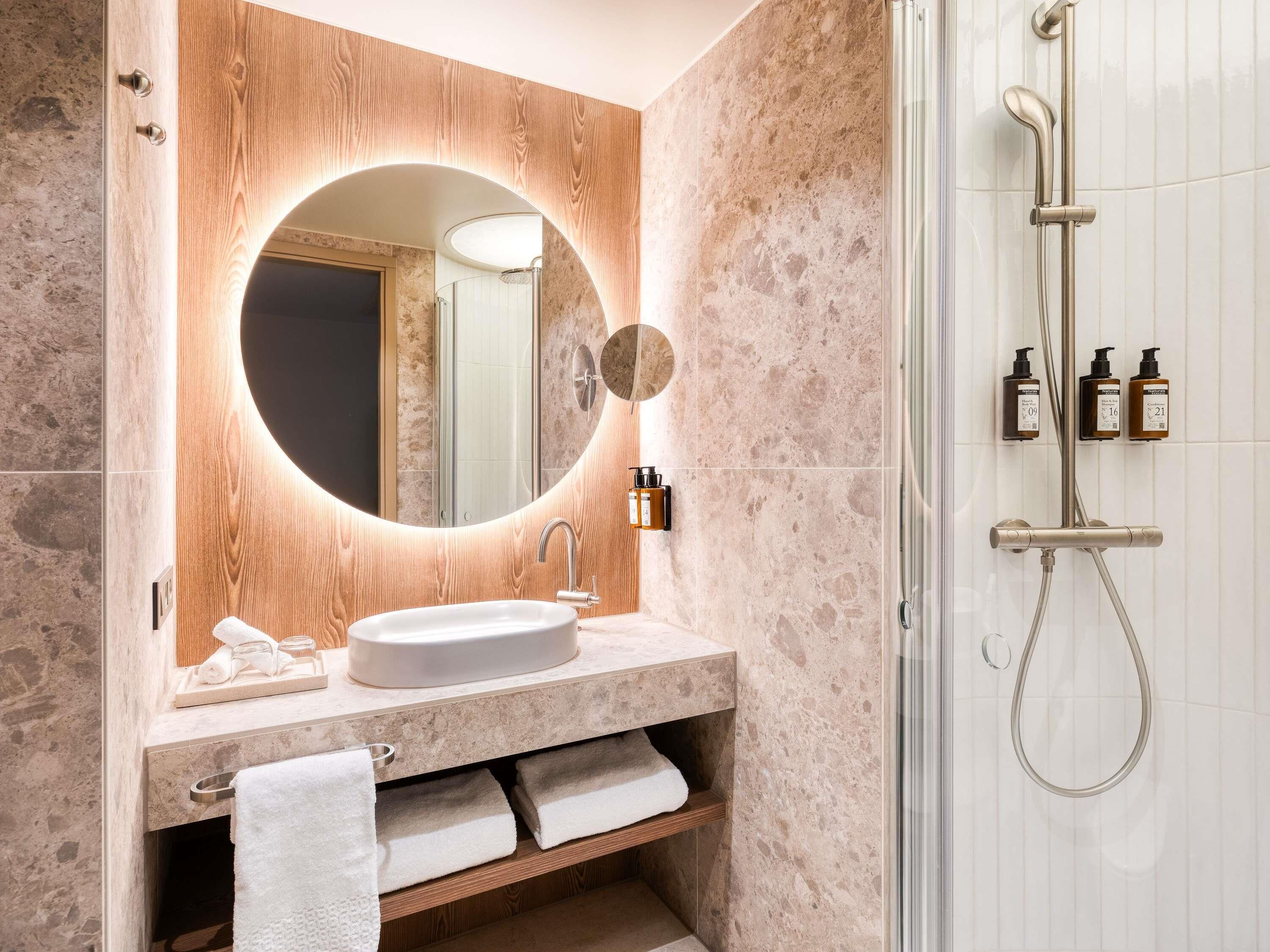 A modern bathroom with a round mirror, a sink, a shower, and toiletries.