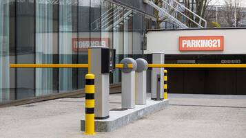 L’entrée d’un parking, avec un potelet jaune et noir à rayures, un panneau indiquant qu’il s’agit d’un parking et une barrière métallique.