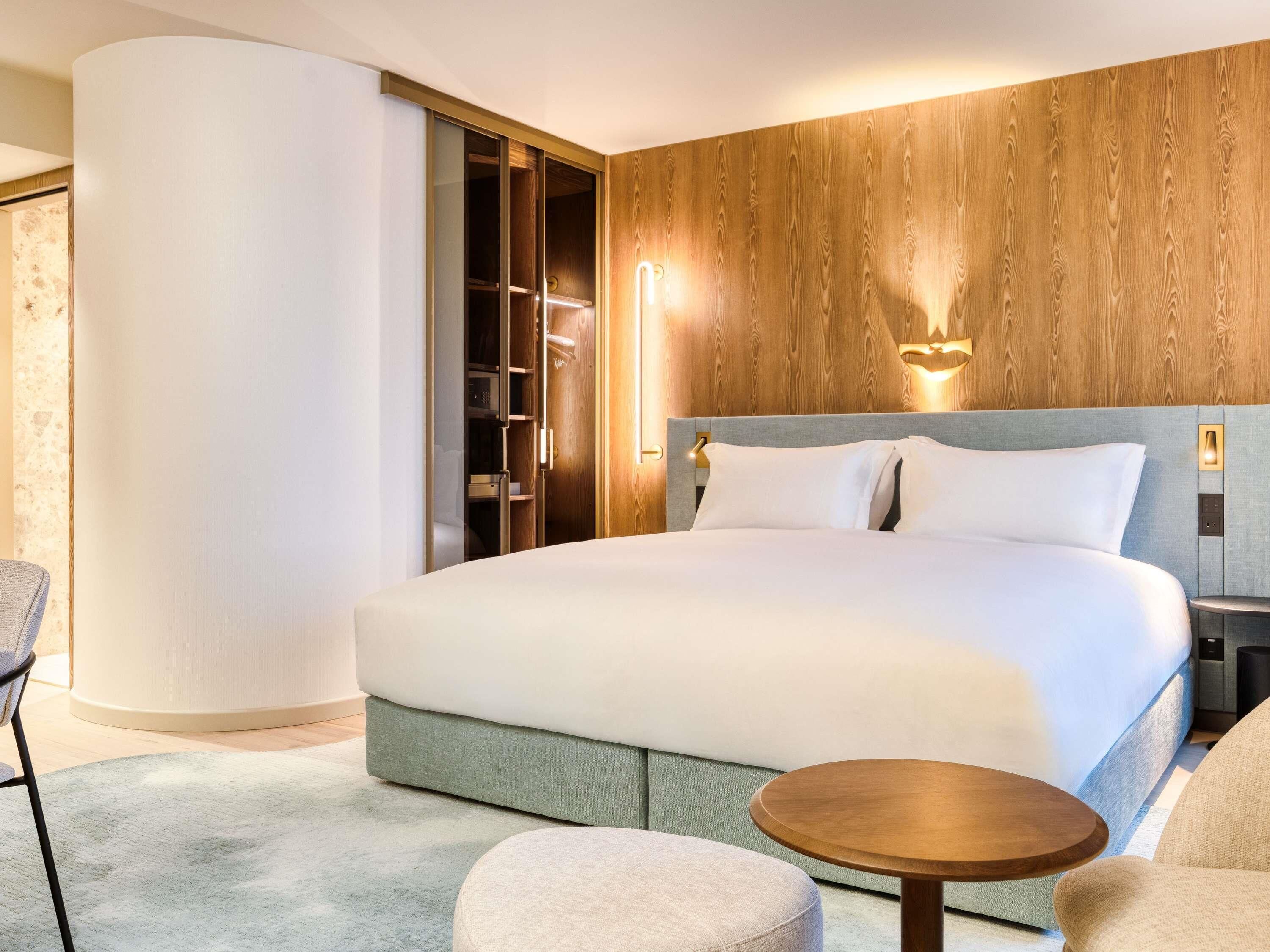 A hotel room with a large bed, a wooden headboard, a round wooden side table, a grey armchair, and a walk-in closet.