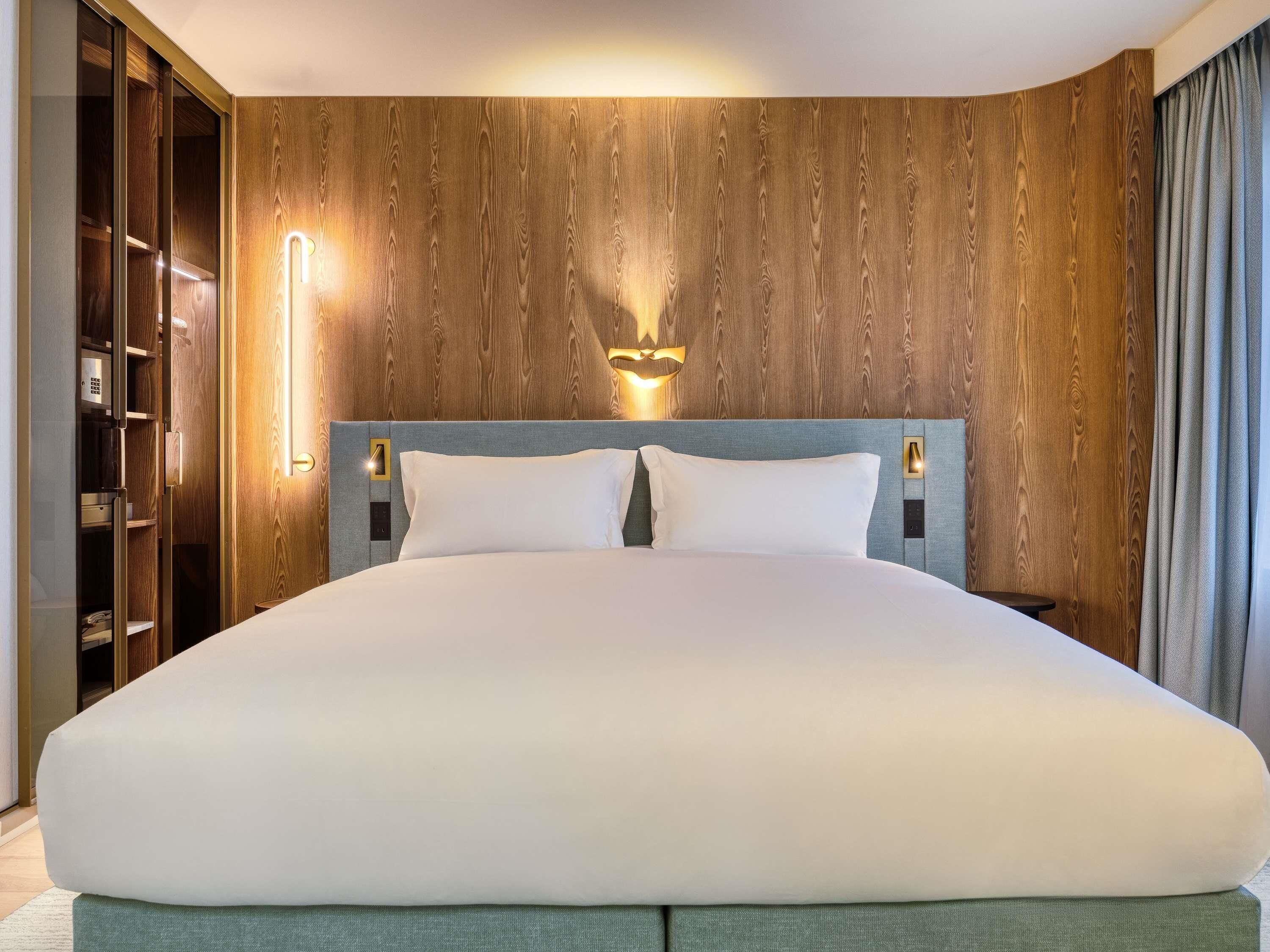 Habitación de hotel con una cama grande, cabecera de madera, armario empotrado y una lámpara fijada a la pared.