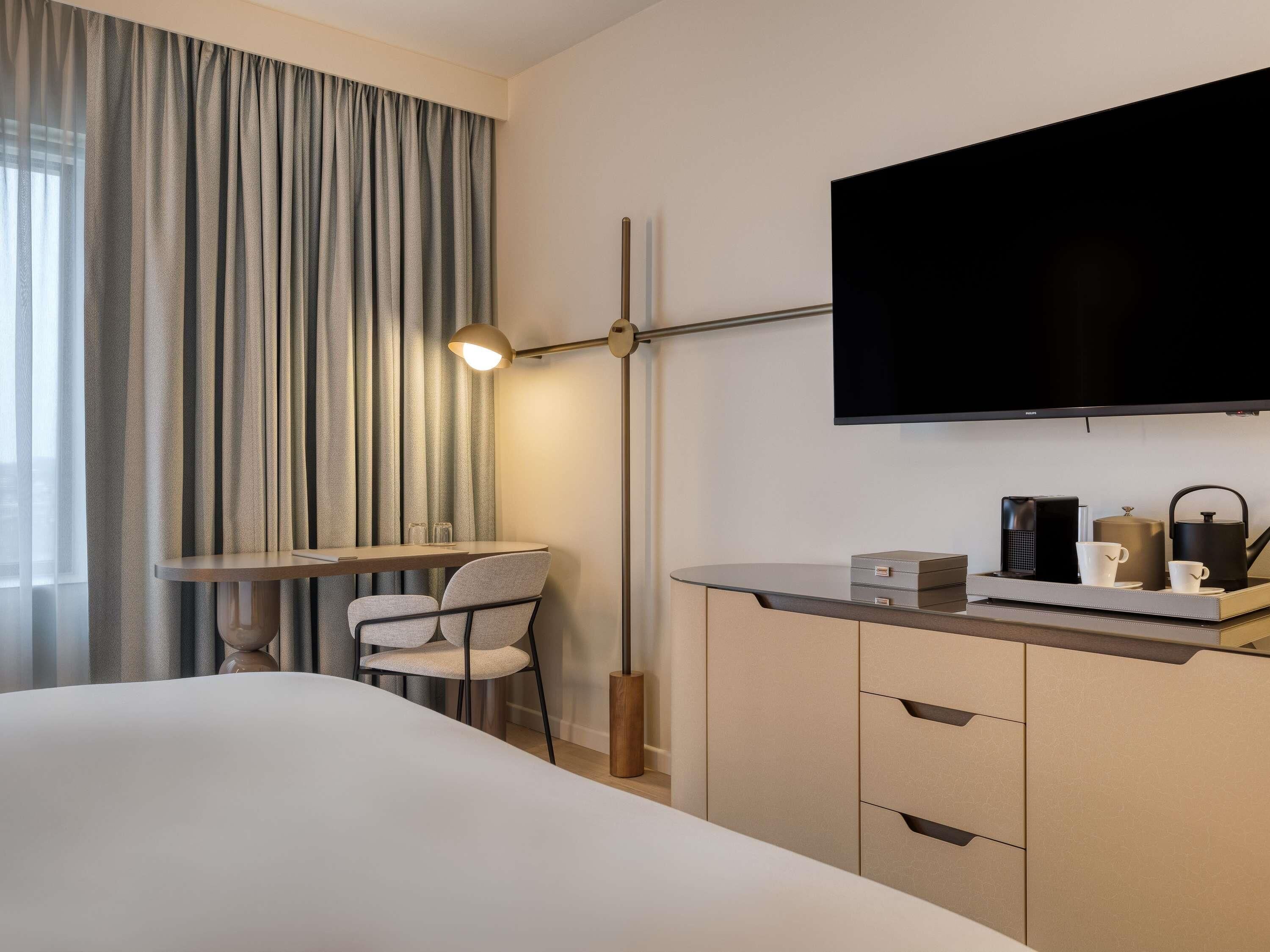 A hotel room with a bed, a desk with a chair, a television, and a minibar.