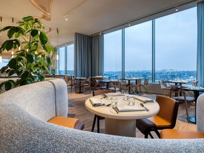 A modern dining area with large windows, a round table set for two, and a large green plant. - Mövenpick hotel Brussels Airport (Zaventem)
