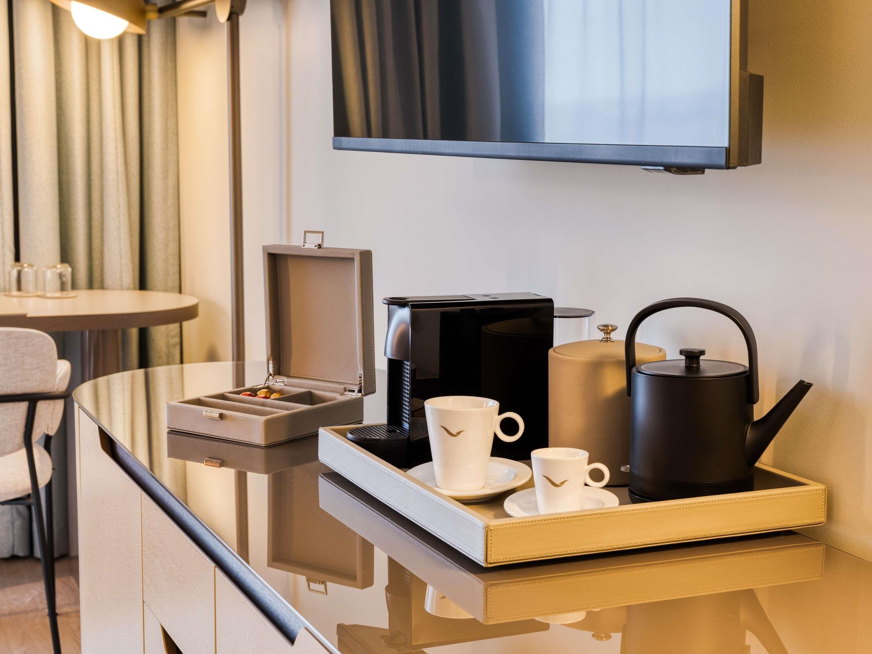 Une chambre d’hôtel avec une table en verre, une machine à café, une bouilloire, un plateau avec des tasses et une boîte de chocolats.