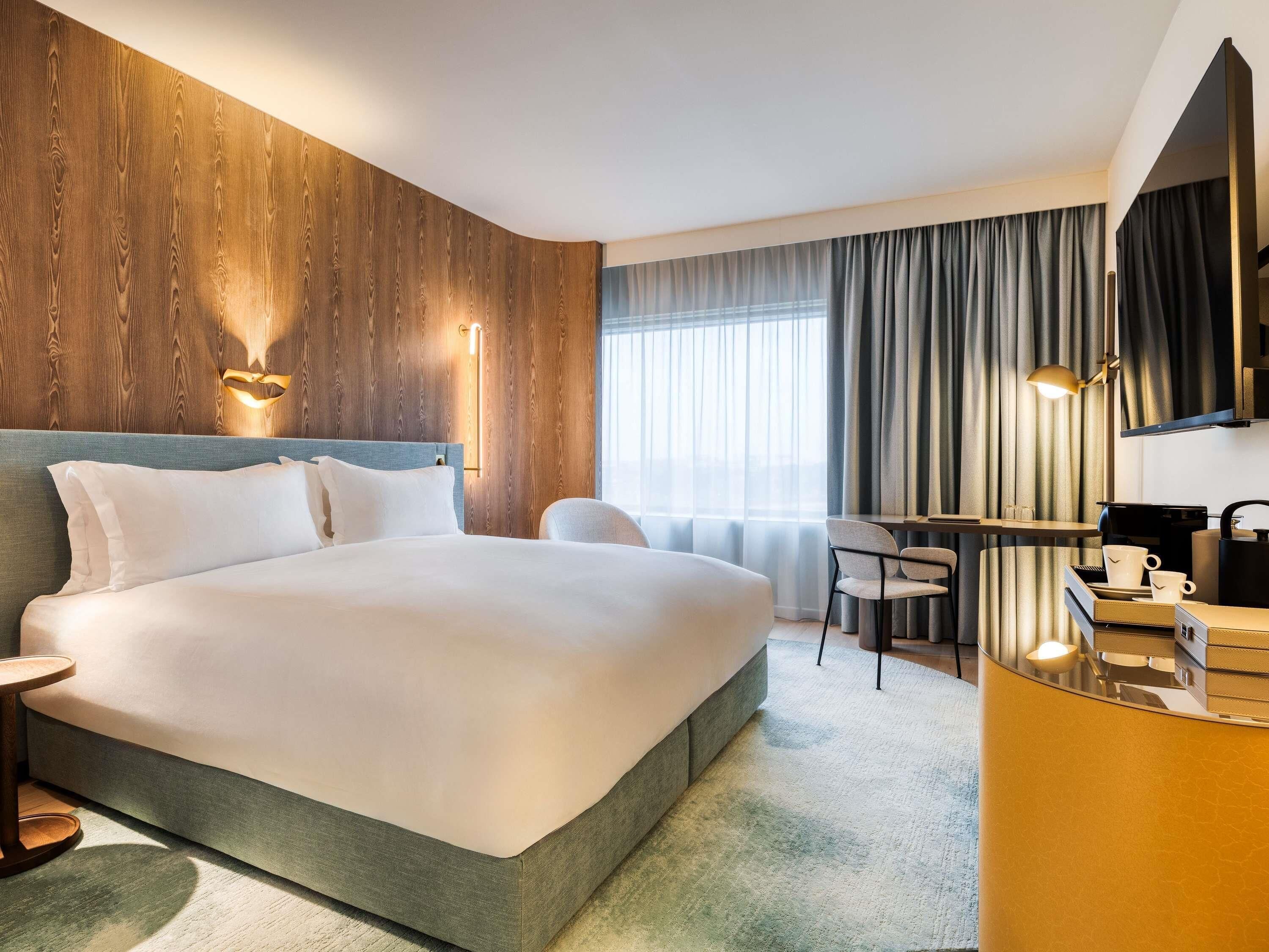 Habitación de hotel con una cama grande, un escritorio con silla, una cafetera y vistas a la ciudad a través de la ventana.