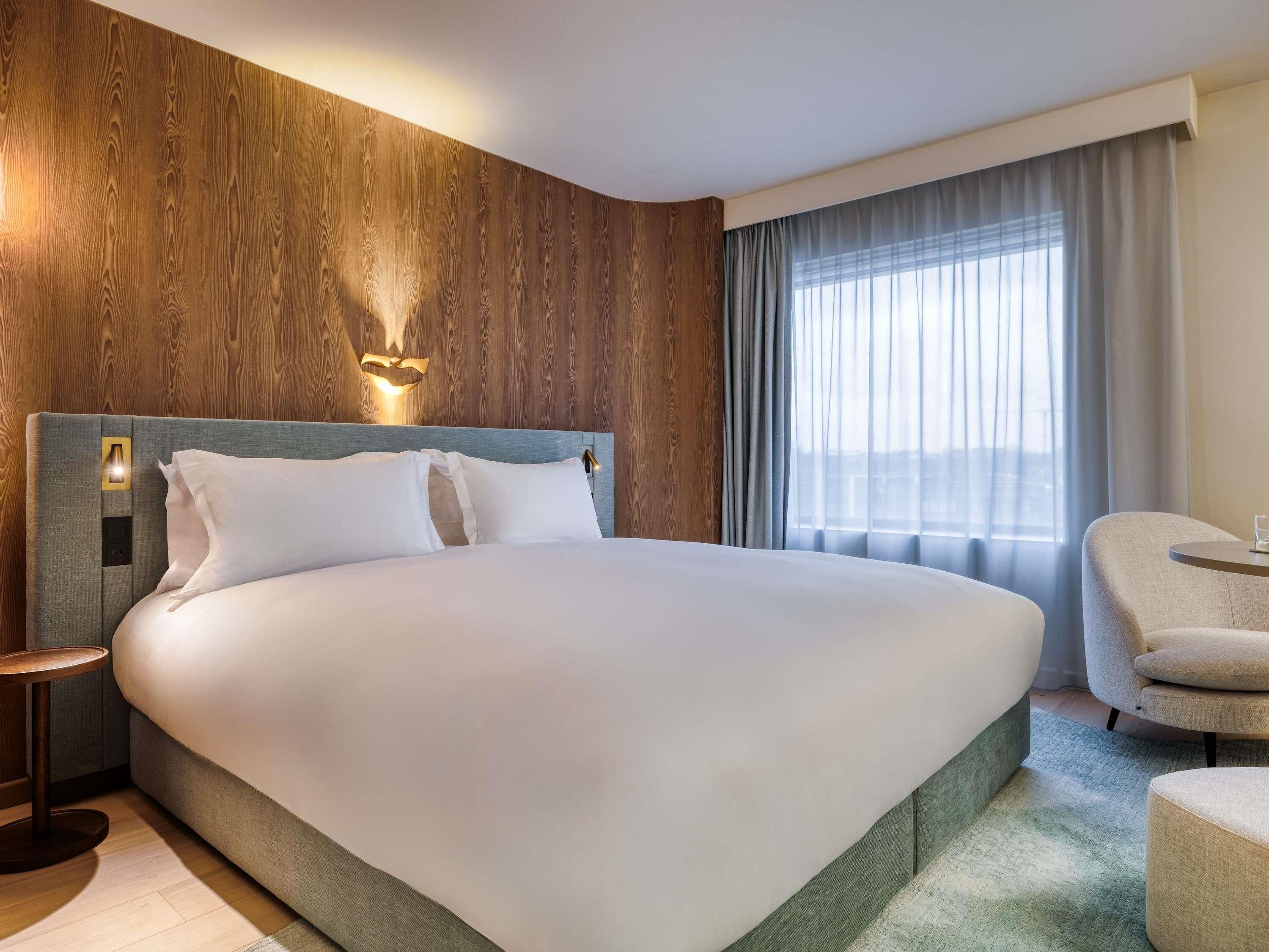 A hotel room with a large bed, a wooden accent wall, a modern headboard, a bedside table, a chair, and a window with curtains.