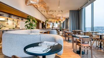 Un restaurante moderno con mesas redondas, una zona de bar y grandes ventanales con vista a la ciudad.