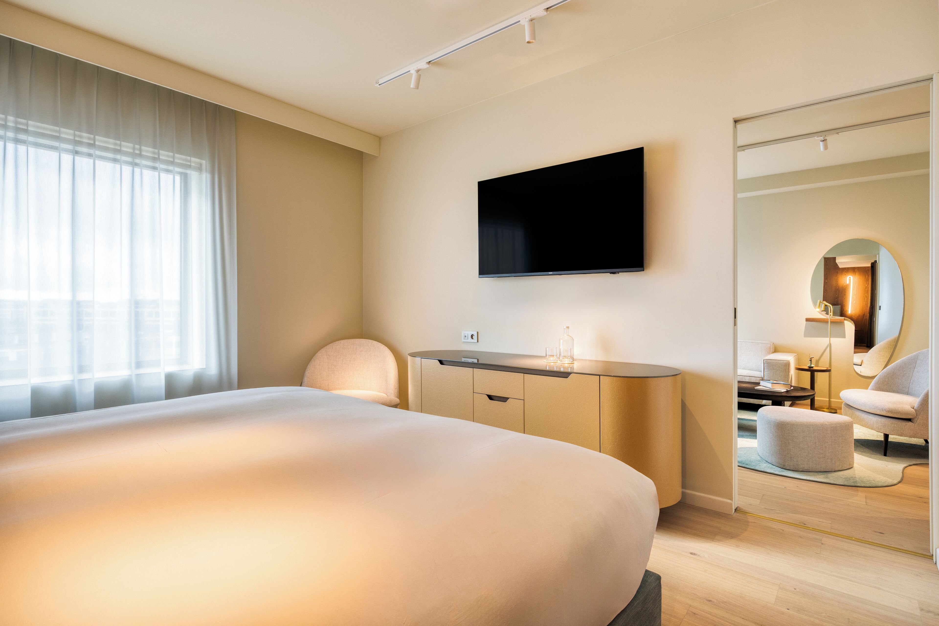 A hotel room with a large bed, a flat-screen TV mounted on the wall, a desk with a lamp, and a mirror.