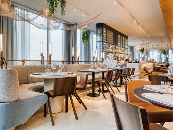 A modern restaurant interior with round tables, cushioned seating, and a bar area. - Mövenpick hotel Brussels Airport (Zaventem)