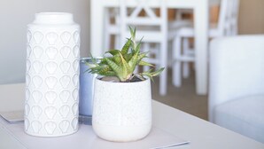 Une table blanche avec une bouteille d’eau blanche et une plante en pot. Au fond, on aperçoit une table à manger blanche et des chaises.
