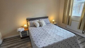 A bedroom with a large bed, two bedside tables with lamps, a window with curtains, and a radiator. - Sittingbourne Serenity - Spacious 4-bed House (Sittingbourne)