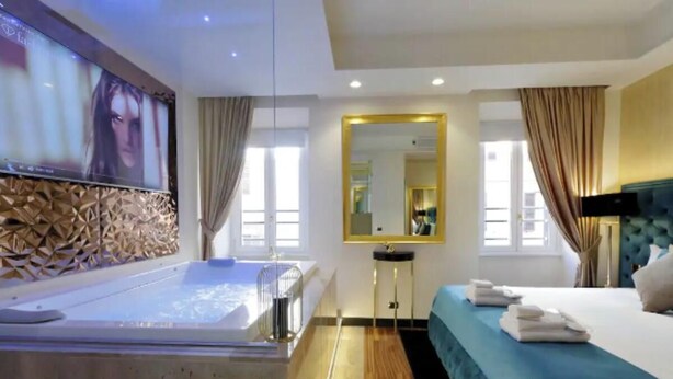 A modern hotel room with a large bathtub, a bed with blue bedding, a mirror, and a television on the wall.