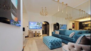 A modern living room with a blue sofa, a tufted ottoman, and a grey armchair. There are two paintings on the wall, a chandelier, and a flat-screen TV.