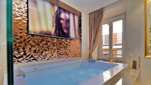 A modern bathroom with a large bathtub, a television mounted on a geometric wall, and a window with curtains.