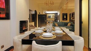 A dining area with a black table set for six, surrounded by white chairs. A chandelier hangs from the ceiling, and a sofa is visible in the background.