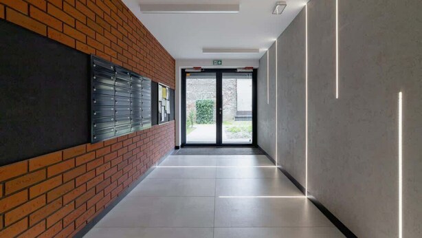 A modern hallway with a brick wall on one side and a plain wall on the other, leading to a glass door with a view of greenery outside.