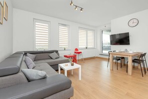 Un salon moderne avec un canapé d’angle gris, une petite table blanche, un fauteuil rouge, une table à manger avec des chaises et un téléviseur fixé au mur.