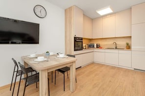 Une cuisine moderne avec des armoires blanches, un four encastré, un micro-ondes et un évier. On y trouve une table à manger avec des chaises et une horloge au mur.