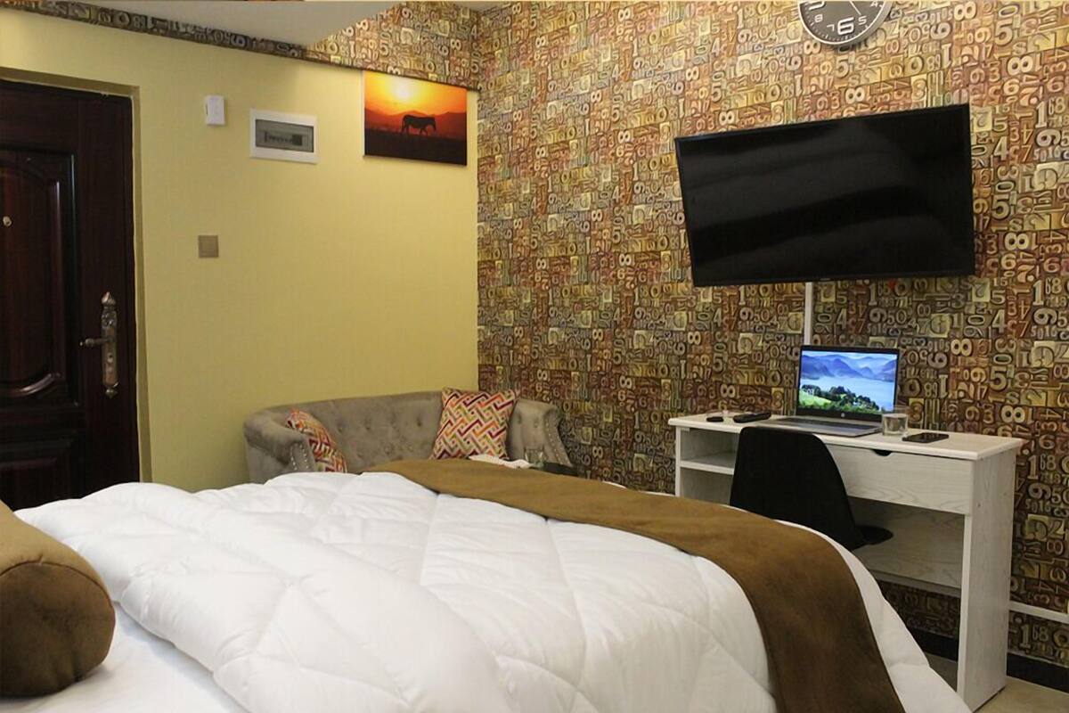 A hotel room with a bed, a desk with a computer, a television, and a patterned wallpaper.