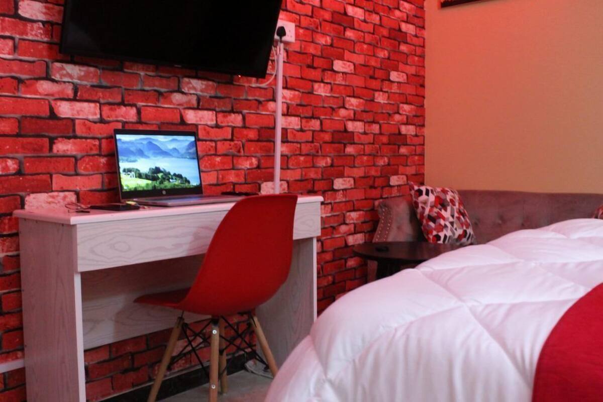 A room with a brick wall, a desk with a computer, a red chair, and a bed with white bedding.