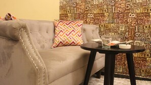 Un canapé à accoudoirs capitonnés avec un coussin à motifs, une petite table ronde avec un verre et un objet blanc, et un mur orné d’un motif numérique.