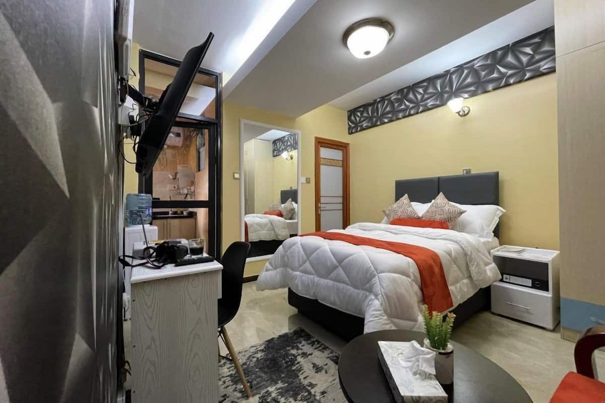 A hotel room with a bed, bedside tables, a desk with a chair, a mirror, and a small plant.