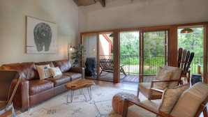A spacious living area with a brown leather sofa, a wooden coffee table, and a patterned rug. There is a large window with a view of greenery outside. - The Burly Bear | AC, Pool, Hot Tub, Golf & Ski (Durango)