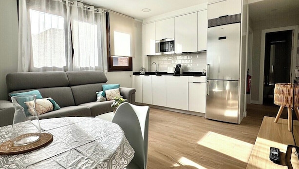 Een moderne keuken met witte kastjes, een roestvrijstalen koelkast en een magnetron. Er is een eethoek met een tafel en stoelen, en een bank in de woonkamer.