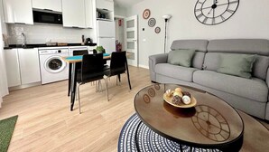 A modern living room with a grey sofa, a round coffee table with decorative items, a dining area with a table and chairs, and a kitchen with white cabinets and appliances. - Vivodomo | Free parking, new, large terrace (Madrid)